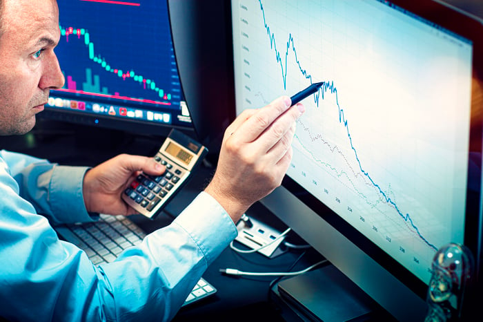 A money manager uses a pen and calculator to check a stock chart displayed on a computer monitor.